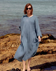 A woman La Bottega di Brunella Linen Dress, long-sleeve blue-gray textured and sunglasses stands barefoot on rocky shoreline with water in the background. A pair of black sandals rests on the rocks beside her.