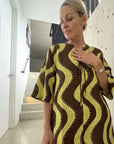 Alemais Linen Dress Worn by woman brown and yellow wavy patterned kaftan dress, standing in a modern interior hallway with concrete walls and stairs.