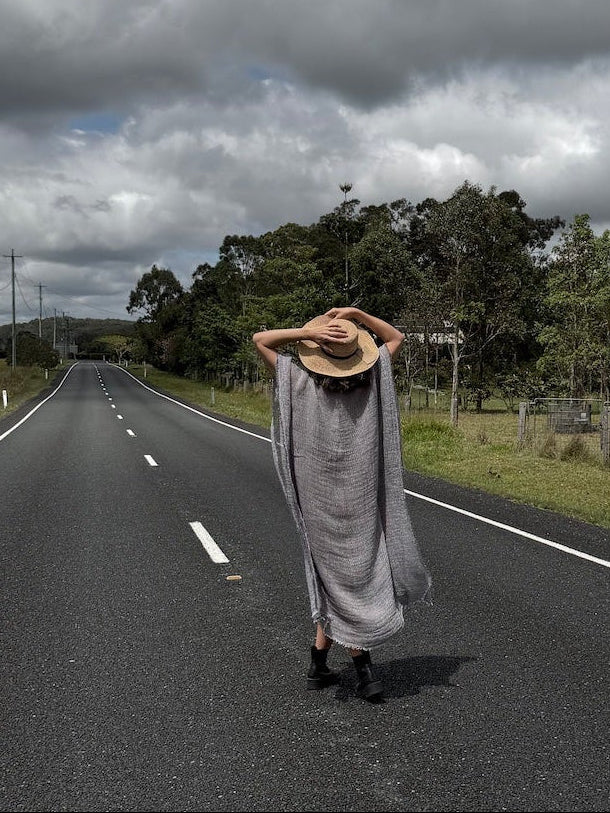 Person in a Estilo Emporio dress and hat walking alone on a long, straight road under a cloudy sky.