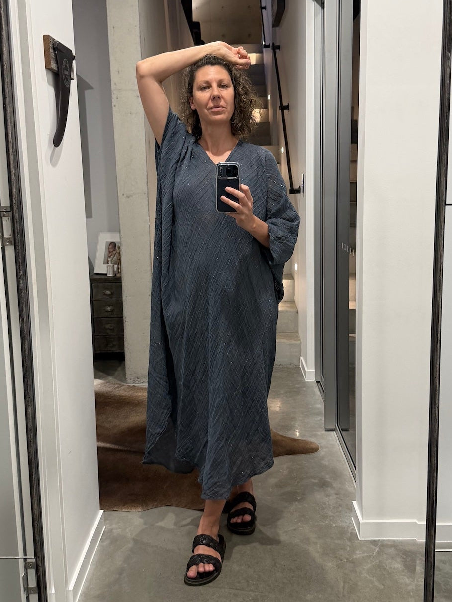 Woman taking a mirror selfie wearing a blue la bottega di brunella linen dress made in italy in a room with white walls and a gray floor.
