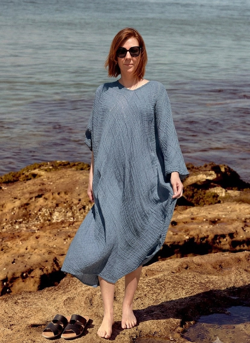 A woman La Bottega di Brunella Linen Dress, long-sleeve blue-gray textured  and sunglasses stands barefoot on rocky shoreline with water in the background. A pair of black sandals rests on the rocks beside her.